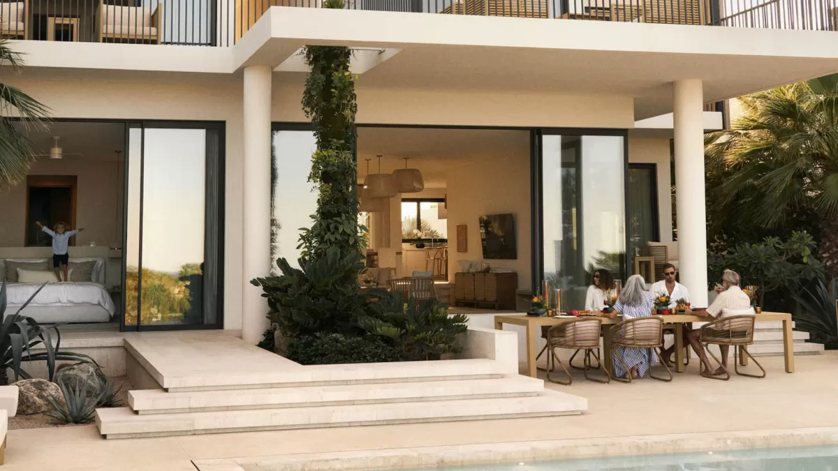 Family dining on a sunlit villa terrace beside a pool, while a child plays on the bed in the open suite behind.