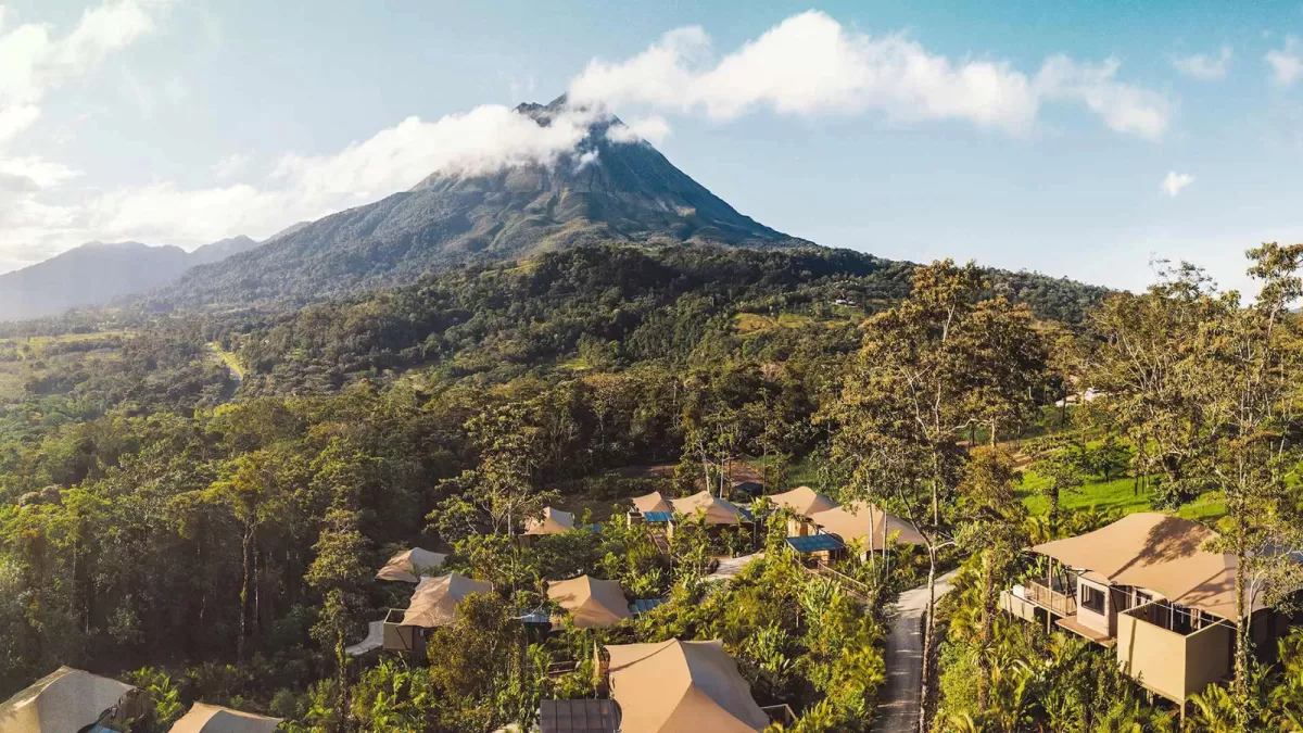 Aerial view of canvas-roofed villas scattered through rainforest below a cloud-capped volcano.